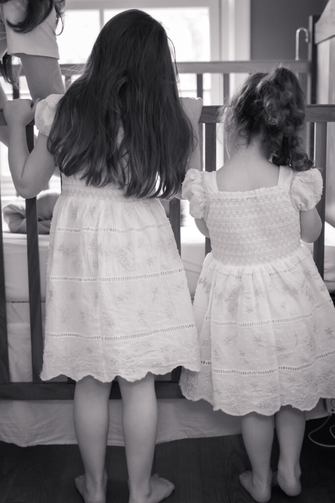 View from behind as two girls peak in on baby brother in crib in black and white.