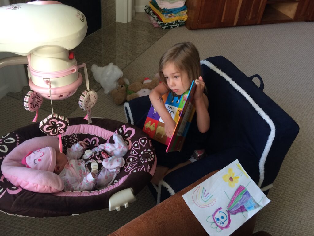 Toddler sibling sitting on chair reading first words book to infant in baby swing.