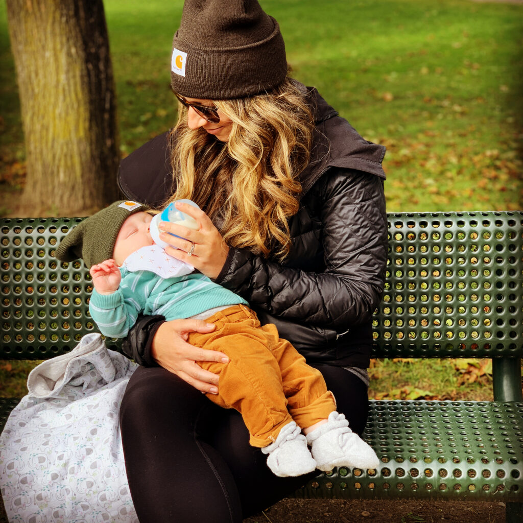 Mother in hat, sun glasses, feeding infant while sitting outside on a park bench.