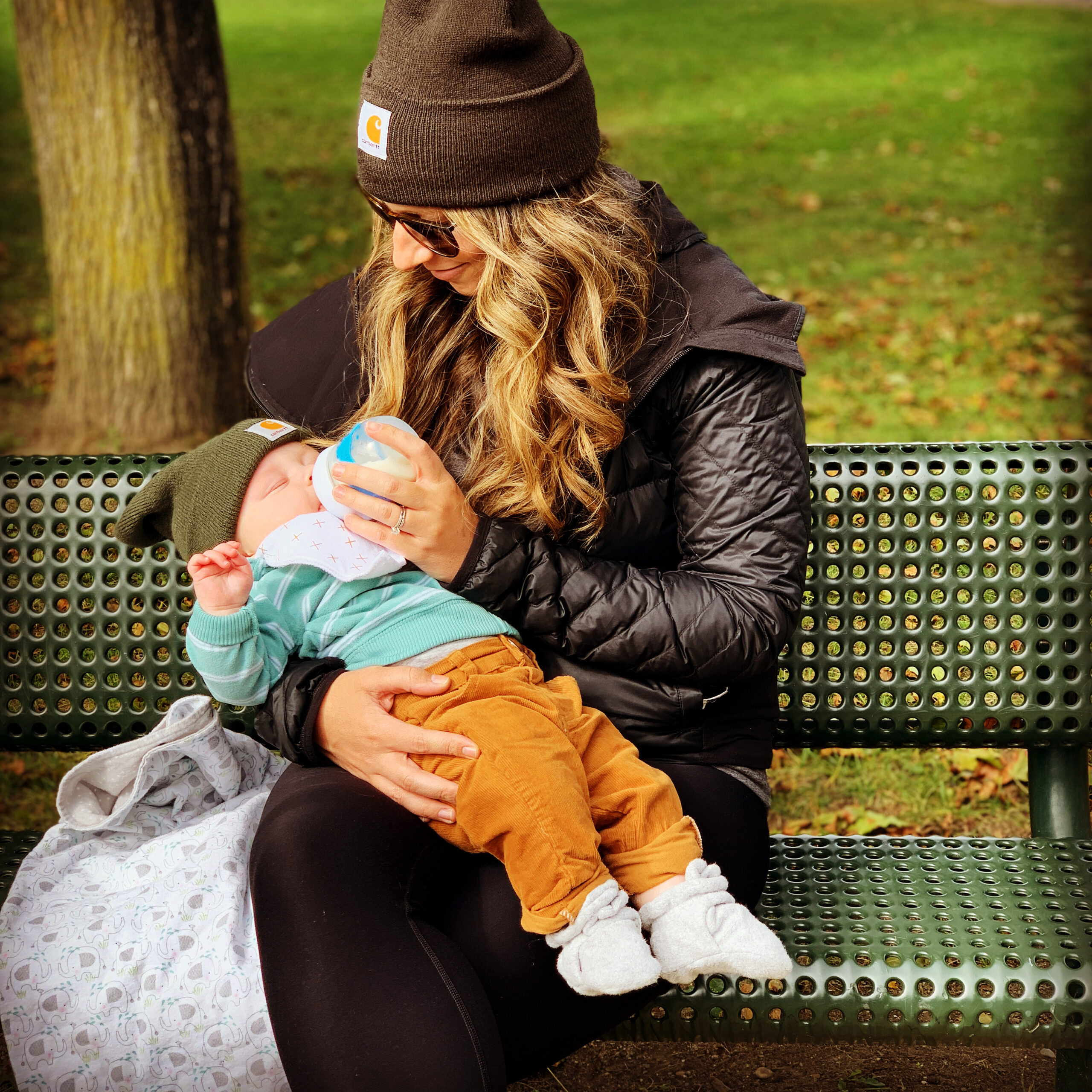 Mother in hat, sun glasses, feeding infant while sitting outside on a park bench.