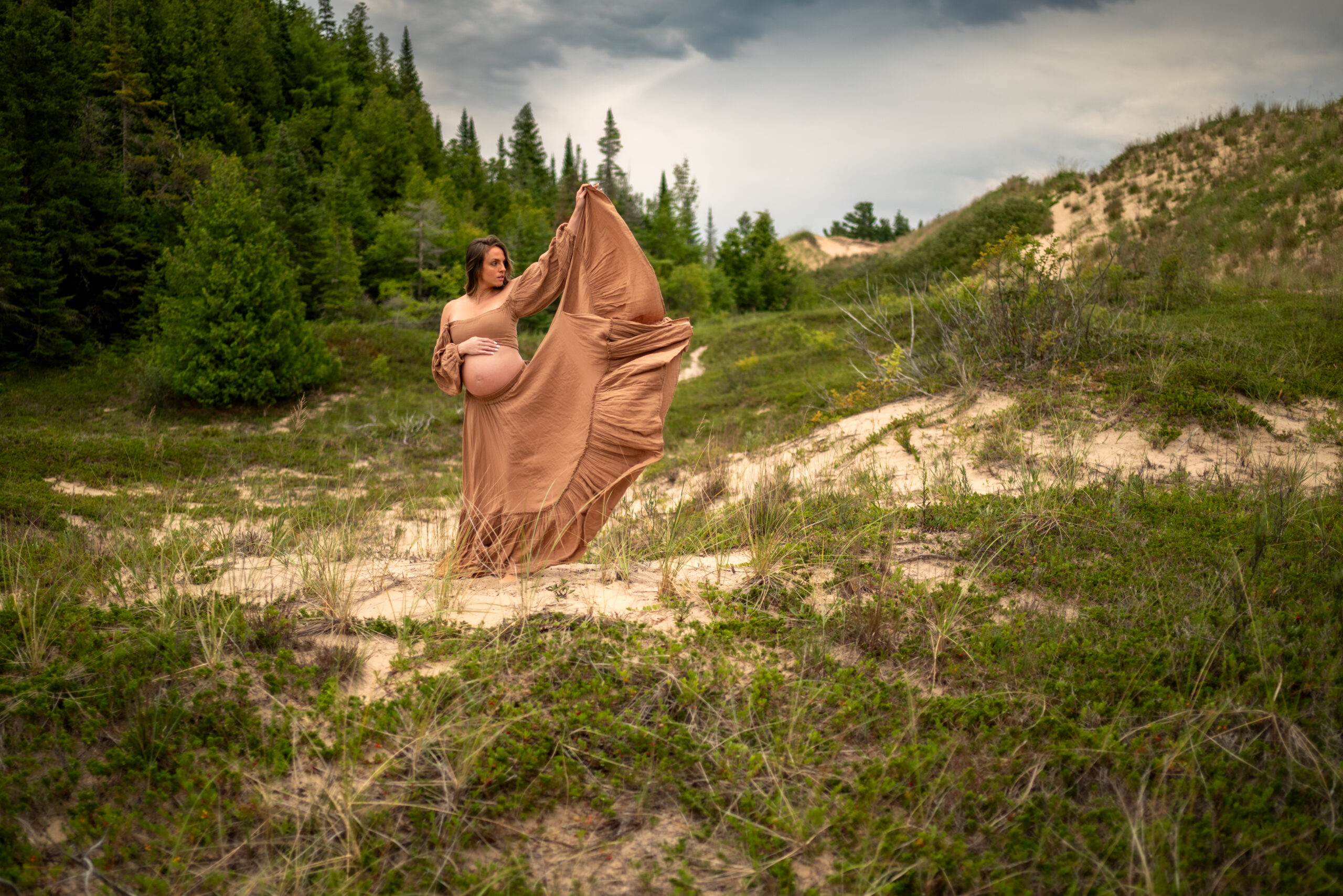 Pregnant woman holding skirt of dress to the sky against dramatic dune background