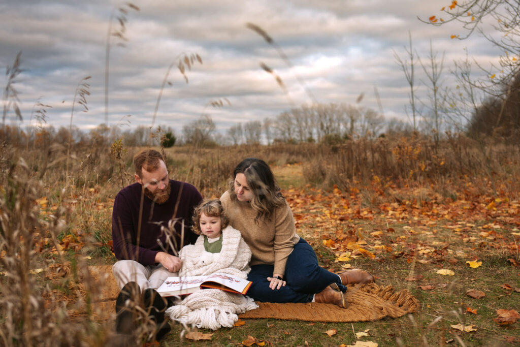 Dad with burgundy sweater and mother with camel sweater cuddle small child on blanket with coordinated green jumper for fall photo.