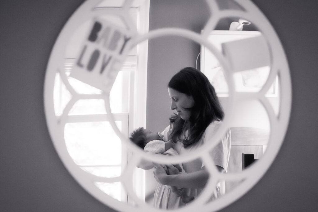A contemplative black and white portrait of a mother and newborn framed in a nursery mirror, bathed in soft natural light.