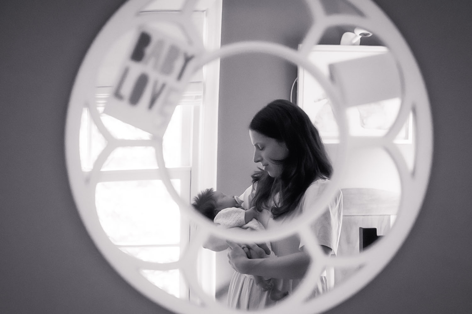 A contemplative black and white portrait of a mother and newborn framed in a nursery mirror, bathed in soft natural light.