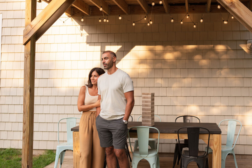 Mother in neutral tank and camel colored pants and father with plain tee and dress shorts watch children play in yard.