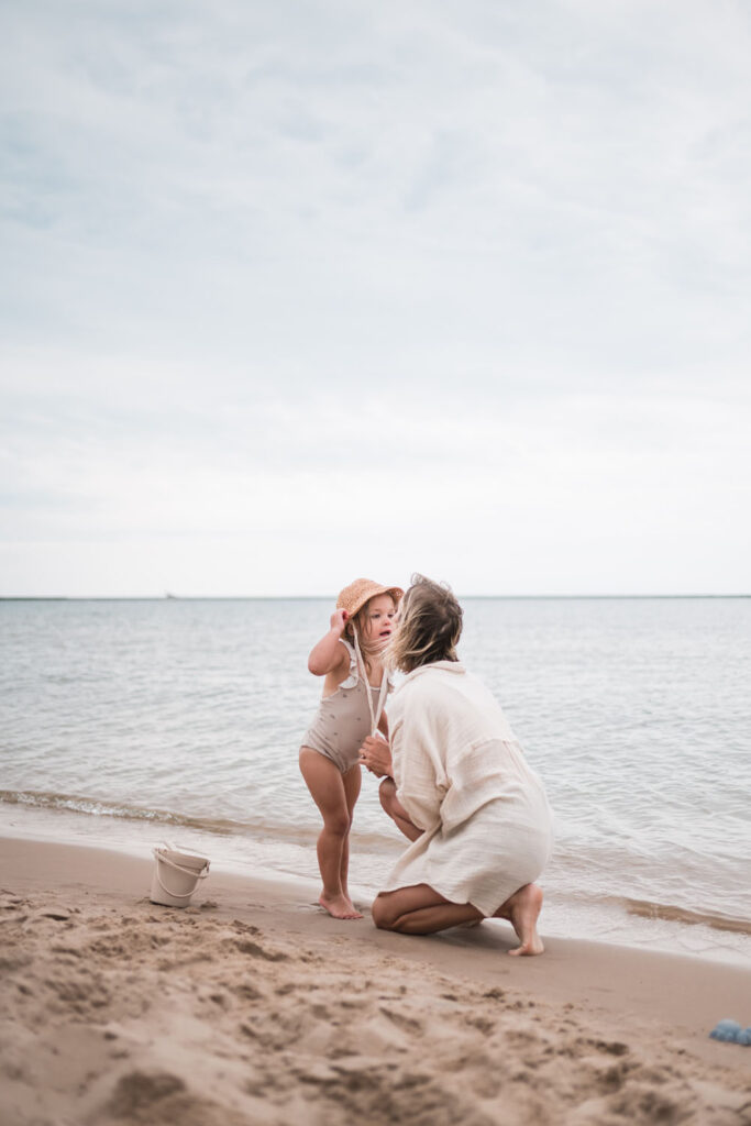 The best places to shop for family photo outfits will feature neutral looks like that found here of a mother in white loose shirt and daughter in swim suite and straw hat share a moment on the shore of lake Michigan. 