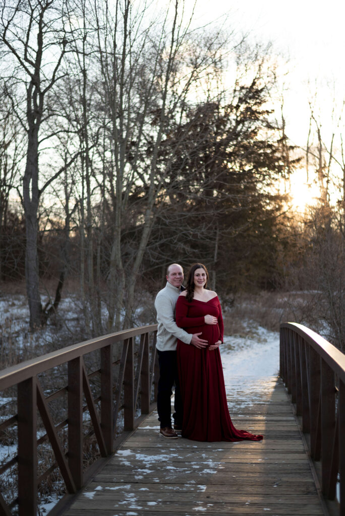Winter maternity photograph showing pregnant woman in long wine-colored dress with her partner dressed in a soft grey-neutral sweater.