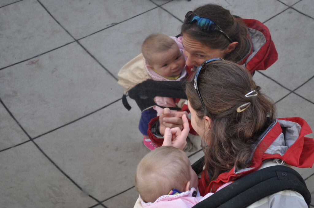mother holding infant in baby carrier while wearing sunglasses, scene reflected
