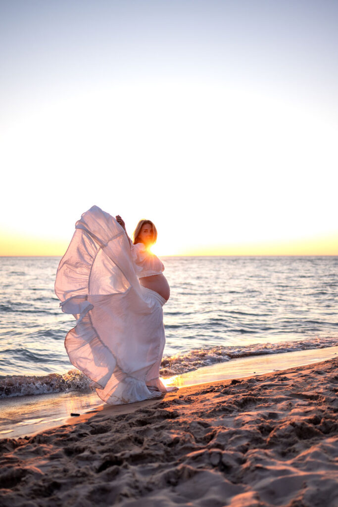 A pregnant woman standing in the setting sun on the beach with sunflare.