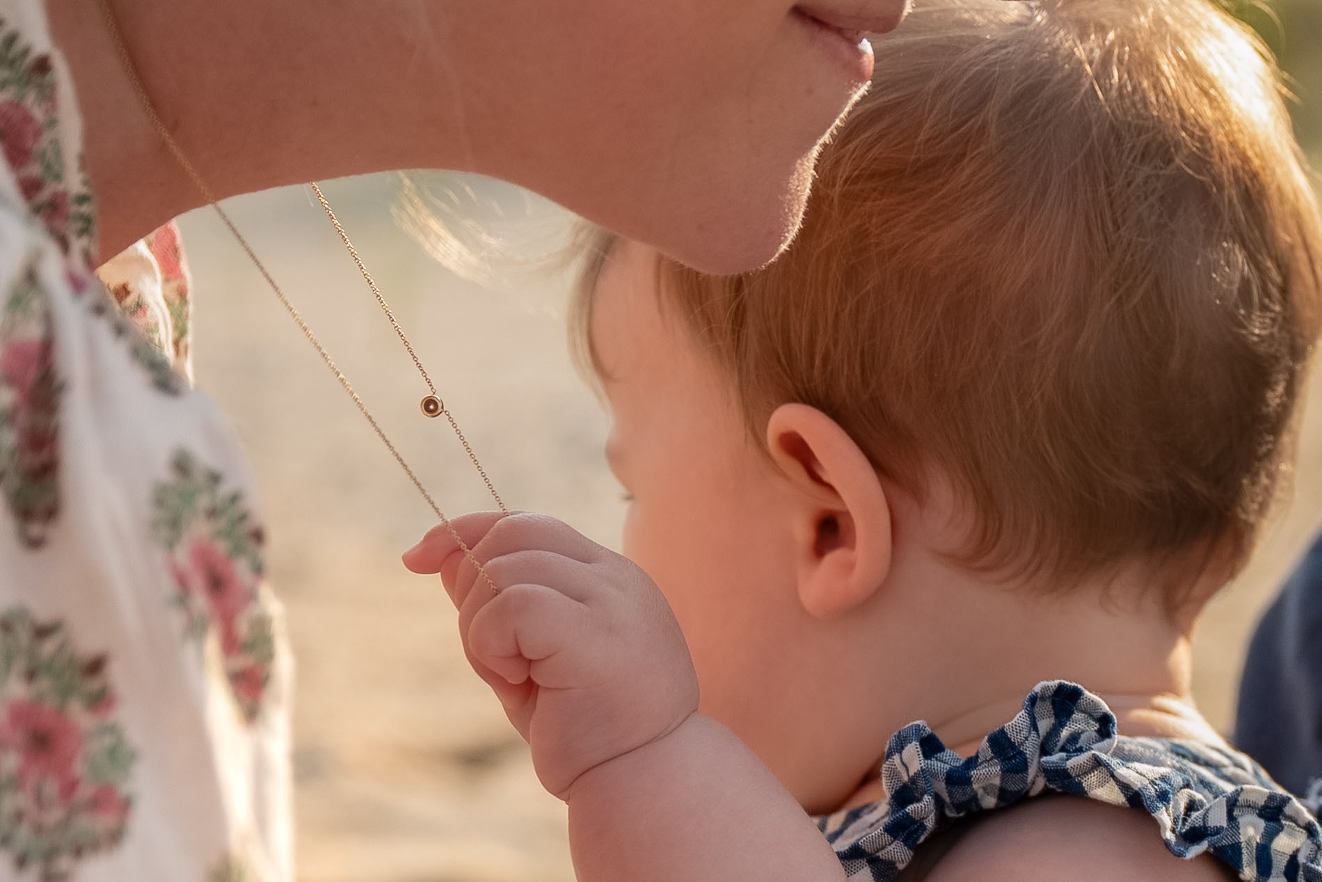 Infant gently tugs mother's necklace in warm morning light.